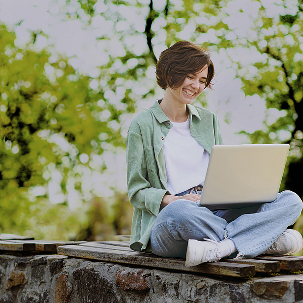 Eine lächelnde Frau sitzt im Freien auf einer Steinmauer und arbeitet an einem Laptop. Sie trägt ein helles Oberteil und eine grüne Jacke. Um sie herum sind grüne Bäume und Pflanzen zu sehen, die eine entspannte Atmosphäre schaffen.
