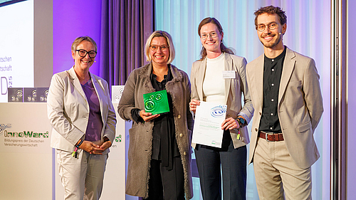 Ein Preisübergabe-Event, bei dem vier Personen auf der Bühne stehen. Eine Frau in der Mitte hält einen Preis und ein Zertifikat. Die anderen drei, darunter eine Frau links mit Brille, lächeln. Im Hintergrund ist ein Banner mit dem Schriftzug „InnoWard“ sichtbar.