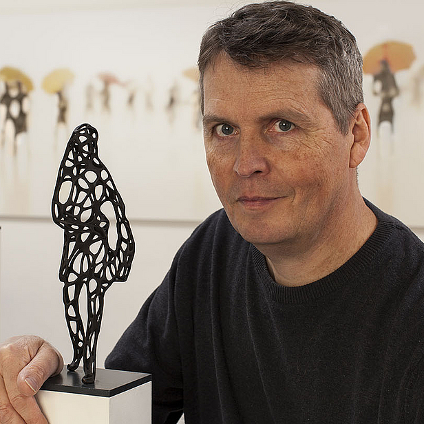 Ein Künstler sitzt neben zwei kunstvollen, abstrakten Skulpturen auf Podesten. Er hat den Blick direkt in die Kamera gerichtet und trägt ein dunkelblaues Shirt. Im Hintergrund sind verschwommene Figuren mit Regenschirmen zu sehen.