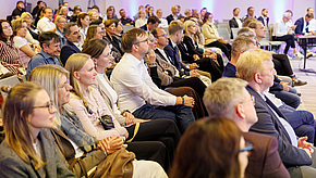 Eine Gruppe von Menschen sitzt in einem gut beleuchteten Raum und hört aufmerksam zu. Sie sind in verschiedene Richtungen ausgerichtet und tragen geschäftliche Kleidung. Die Atmosphäre ist konzentriert und professionell, wobei einige Personen Notizen machen oder sich unterhalten.