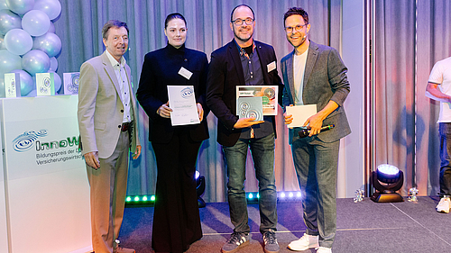 Eine Gruppe von vier Personen steht auf einer Bühne, umgeben von festlicher Dekoration. Sie halten Urkunden in den Händen, die auf eine Auszeichnung hinweisen. Der Hintergrund zeigt eine moderne Veranstaltung mit Lichtern und einem Logo, das den Namen „Innow“ trägt.