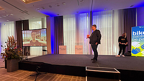 Eine Präsentation findet auf einer Bühne statt. Ein Mann spricht, während ein Bildschirm neben ihm eine Person zeigt, die zugeschaltet ist. Im Hintergrund sind hohe Fenster und ein Banner mit dem Text „bikö – be part of it“.