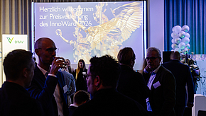 Eine Gruppe von Personen steht in einem festlichen Raum. Im Hintergrund ist eine große Leinwand mit der Aufschrift „Herzlich willkommen zur Preisverleihung des InnoWard 2026“ zu sehen. Die Atmosphäre wirkt elegant und einladend.