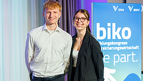 Zwei junge Menschen stehen lächelnd vor einem Plakat mit dem Schriftzug "biko" und Informationen zu einem Bildungs- und Versicherungskongress. Der Mann trägt ein helles Hemd, die Frau eine schwarze Jacke. Beide schauen direkt in die Kamera.