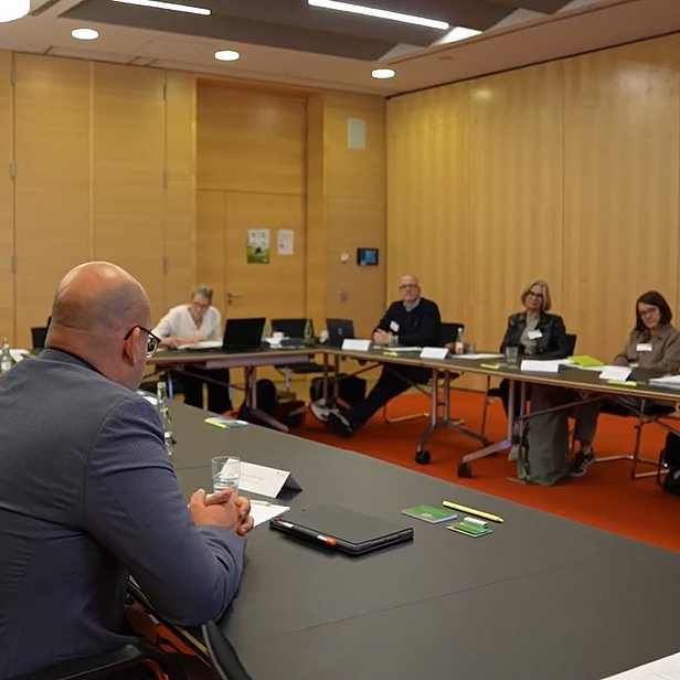 Gruppenversammlung in einem Konferenzraum. Teilnehmer sitzen an einem großen Tisch und diskutieren. Im Vordergrund ist ein Mann mit Glatze zu sehen, der gerade spricht. Die Umgebung ist modern mit Holzvertäfelung und einfacher Tischdekoration.