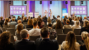 Ein Publikum sitzt in einem Konferenzraum und blickt auf eine Präsentation. Auf der Bühne stehen zwei Personen, die sprechen. Im Hintergrund sind Grafiken und Bilder zu sehen, die Informationen darstellen. Die Atmosphäre ist professionell und gleichzeitig interaktiv.