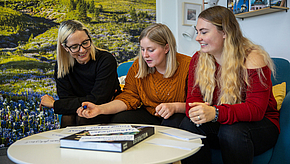 Drei Frauen sitzen um einen Tisch und diskutieren lebhaft. Eine Frau mit blonden Haaren trägt eine schwarze Bluse, die zweite hat mittellange blonde Haare und ein orangefarbenes Oberteil, während die dritte lange blonde Haare und ein rotes Oberteil hat. Auf dem Tisch liegt ein Buch.