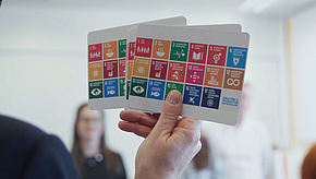 Eine Hand hält mehrere Karten mit ikonischen Darstellungen der UN-Nachhaltigkeitsziele. Die Karten sind bunt und zeigen verschiedene Themen wie Gesundheit, Bildung und Umwelt. Im Hintergrund sind unscharf Menschen zu sehen, die an einer Diskussion teilnehmen.