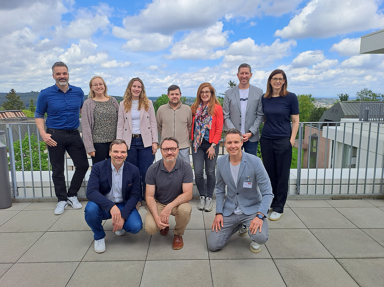Eine Gruppe von zwölf Personen steht und sitzt auf einer Terrasse. Im Hintergrund sind Wolken und Gebäude sichtbar. Die Anwesenden lächeln und tragen legere Kleidung. Einige sind in herausragenden Farben gekleidet, was ein geselliges und freundliches Ambiente vermittelt.