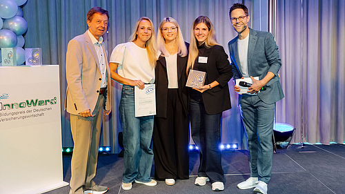 Vier Personen stehen auf einer Bühne, lächelnd und in eleganter Kleidung. Sie halten Auszeichnungen in der Hand. Im Hintergrund ist eine Präsentation und dekorative Beleuchtung sichtbar. Es scheint eine Preisverleihung oder Ehrung zu sein.