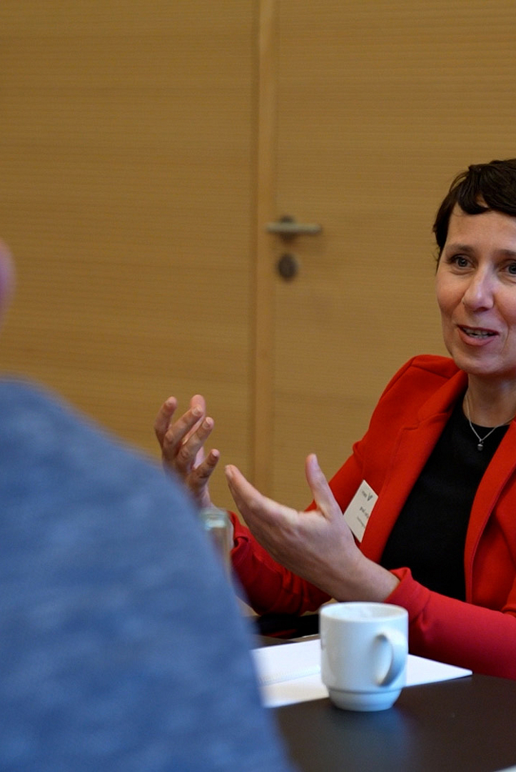 Eine Frau in einem roten Blazer sitzt an einem Tisch und gestikuliert beim Sprechen. Vor ihr steht eine Tasse. Im Hintergrund sind unscharfe Personen zu sehen, die ihr zuhören. Die Szene vermittelt eine Diskussion oder ein Gespräch.