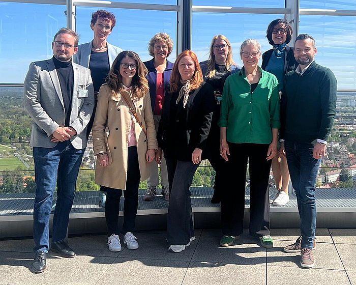 Eine Gruppe von neun Personen steht in zwei Reihen vor großen Fenstern mit Blick auf eine grüne Landschaft. Die meisten tragen formelle Kleidung. Sie lächeln und wirken freundlich, während sie in die Kamera schauen.