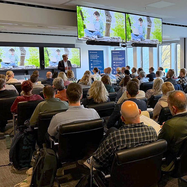 Ein Vortrag vor einem Publikum in einem hellen Raum. Der Redner steht auf der Bühne, während große Bildschirme hinter ihm Präsentationen zeigen. Die Zuhörer sitzen in einem Seminarraum und hören aufmerksam zu.