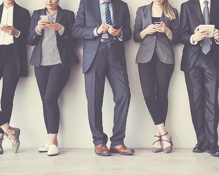 Sechs Personen in formeller Kleidung stehen nebeneinander und nutzen verschiedene digitale Geräte, darunter Smartphones und Tablets. Die Gruppe zeigt eine Mischung aus Männern und Frauen, die mit ihren Geräten beschäftigt sind, ohne in die Kamera zu schauen.