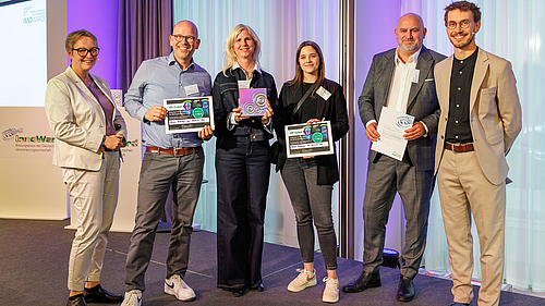 Sechs Personen stehen auf einer Bühne und halten Auszeichnungen in den Händen. Sie lächeln und sind in formeller Kleidung gekleidet. Der Hintergrund ist hell und professionell, was den feierlichen Anlass unterstreicht. Es handelt sich um eine Preisverleihung.