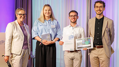 Vier Personen stehen auf einer Bühne bei einer Preisverleihung. Zwei Empfänger halten Urkunden und Zertifikate. Alle Personen lächeln und tragen formelle Kleidung. Der Hintergrund ist in sanften Farben, die die festliche Atmosphäre unterstreichen.