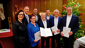 Eine Gruppe von sieben Personen steht zusammen, einige halten Urkunden oder Dokumente. Im Hintergrund steht ein Weihnachtsbaum. Die Menschen lächeln und scheinen an einem feierlichen Anlass teilzunehmen.