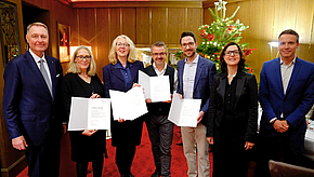 Eine Gruppe von sechs Personen steht in einem festlichen Raum, lächelnd, und hält jeweils Urkunden oder Dokumente in den Händen. Im Hintergrund ist ein dekorierter Weihnachtsbaum zu sehen. Die Atmosphäre wirkt feierlich und herzlich.