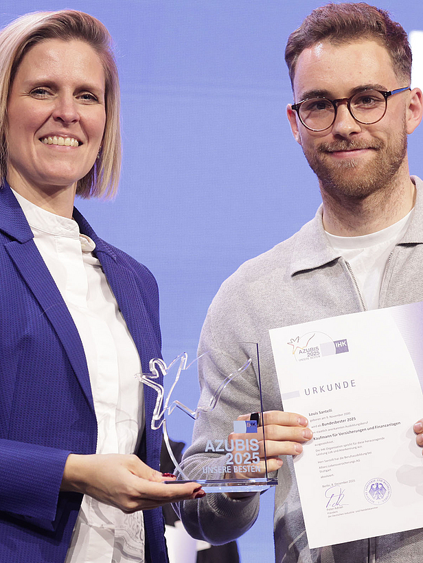 Eine Frau überreicht einem Mann einen Preis und eine Urkunde auf einer Bühne. Beide lächeln in die Kamera. Die Frau trägt einen blauen Blazer, der Mann ein graues Sweatshirt und eine Brille. Im Hintergrund sind weitere Personen sichtbar.