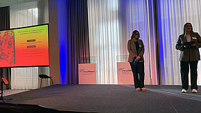 Zwei Personen stehen auf einer Bühne und präsentieren Informationen. Im Hintergrund ist ein Bildschirm mit farbigen Grafiken und Text zu sehen. Die Umgebung ist modern, mit großen Fenstern und einem professionellen Setting.