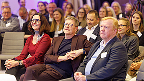 Eine Gruppe von Menschen sitzt in einem Veranstaltungsraum und hört aufmerksam zu. Im Vordergrund sind drei Personen zu sehen: eine Frau in rotem Oberteil, ein Mann mit Brille und ein weiterer Mann in einem dunklen Blazer. Alle wirken interessiert und engagiert.