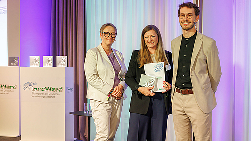 Drei Personen stehen auf einer Bühne, während eine Frau zwischen ihnen ein Zertifikat hält. Im Hintergrund sind Podien mit dem Logo "InnoWards" zu sehen. Die Atmosphäre ist feierlich und professionell, was auf eine Preisverleihung oder Auszeichnung hinweist.