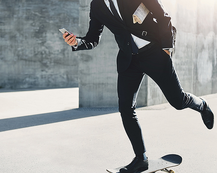 Ein Mann in einem Anzug fährt auf einem Skateboard. Er hält ein Handy in der Hand und scheint in Eile zu sein. Der Hintergrund besteht aus grauem Beton, was einen modernen, urbanen Eindruck vermittelt.