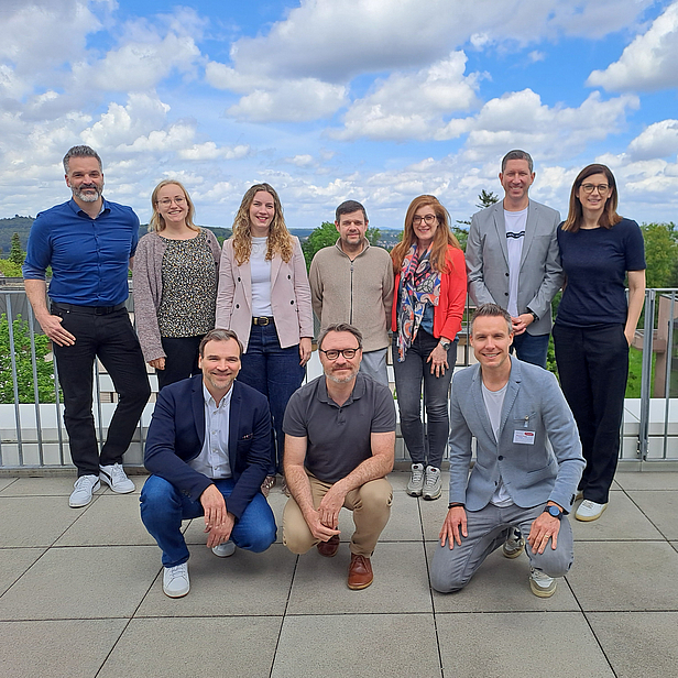 Die Abbildung zeigt eine Gruppe von acht Personen, die auf einem Balkon oder einer Terrasse stehen. Sie sind lässig gekleidet und posieren freundlich. Im Hintergrund sind eine grüne Landschaft und ein bewölkter Himmel zu sehen.