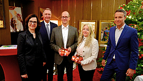 Fünf Personen stehen gruppiert vor einem Weihnachtsbaum. Sie halten kleine, eingepackte Geschenke in den Händen. Die Atmosphäre wirkt festlich und freundlich, was auf eine besondere Anlässe hindeutet.