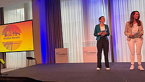 Zwei Frauen stehen auf einer Bühne und präsentieren ein Thema mit dem Titel "Hidden Heroes". Im Hintergrund ist eine große Bildschirmprojektion sichtbar, die das Logo und den Titel der Präsentation zeigt. Der Raum ist hell und modern gestaltet.