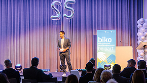 Ein Mann steht auf einer Bühne vor einem Publikum. Er hält ein Mikrofon und spricht. Im Hintergrund sind Luftballons und ein Banner mit der Aufschrift "biko" und "be part." sichtbar. Die Atmosphäre wirkt festlich und professionell.