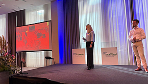 Eine Frau und ein Mann stehen auf einer Bühne und präsentieren vor einer Leinwand. Die Leinwand zeigt eine lebendige, rote Grafik mit verschiedenen Figuren. Im Hintergrund sind Vorhänge und einige Pflanzen sichtbar.