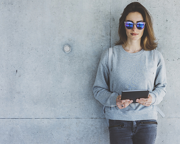 Eine junge Frau mit Sonnenbrille steht lässig an einer Betonwand. Sie trägt einen grauen Pullover und hält ein Tablet in den Händen. Ihr Blick ist auf das Gerät gerichtet, während sie entspannt und modern wirkt.