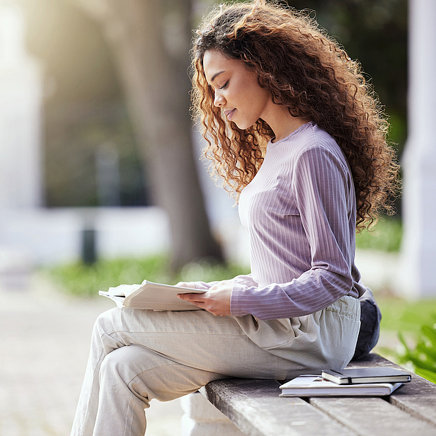 Eine junge Frau mit lockigem Haar sitzt auf einer Bank und liest ein Buch. Sie trägt ein langärmliges Oberteil und eine bequeme Hose. Um sie herum ist eine grüne Umgebung, die eine ruhige, entspannte Atmosphäre schafft.