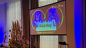 Ein Bildschirm zeigt den Schriftzug "RockRäp" mit dem Untertitel "Wie Bildung Generationen verbindet." Im Hintergrund sind stilisierte Abbildungen von zwei Personen zu sehen. Die Präsentation steht im Kontext eines Bildungs-Events. 