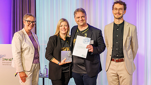 Vier Personen stehen auf einer Bühne. Zwei Personen halten eine Auszeichnung und ein Zertifikat. Im Hintergrund sind dekorative Elemente sichtbar. Alle scheinen lächelnd und stolz zu sein. Die Atmosphäre wirkt feierlich.