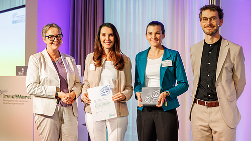 Vier Personen stehen auf einer Bühne und halten Preise in den Händen. Eine Frau mit einer Urkunde, zwei weitere Frauen mit einem Preis und ein Mann, der ebenfalls einen Preis hält. Der Hintergrund ist hell und elegant, was eine feierliche Atmosphäre schafft.