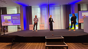 Drei Personen stehen auf einer Bühne bei einer Veranstaltung. Links und rechts sind Projektionsleinwände mit Präsentationen zu sehen. Die Atmosphäre ist freundlich und professionell, mit blauen Lichtern, die den Raum illuminieren.