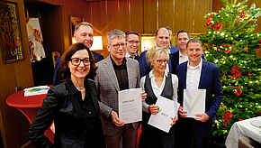 Eine Gruppe von zehn Personen steht in einem festlich dekorierten Raum, umgeben von einem Weihnachtsbaum. Sie halten Urkunden und Dokumente in den Händen. Die Atmosphäre wirkt feierlich und herzlich.