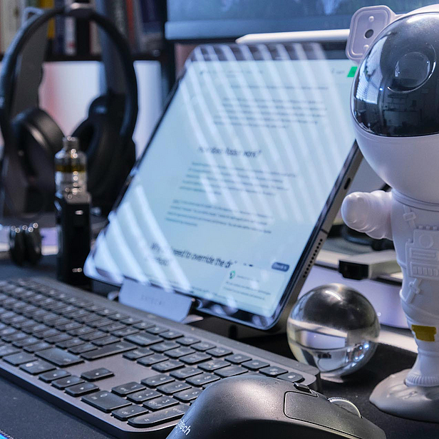 Auf einem Schreibtisch stehen ein Astronauten-Figur, eine Tastatur, ein Tablet mit einem Text und eine Glaskugel. Im Hintergrund sind Kopfhörer und ein Dampfgerät zu sehen. Die Szene vermittelt eine kreative und technologische Arbeitsumgebung.