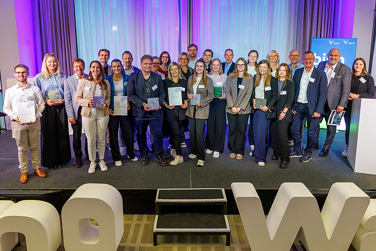 Eine Gruppe von Menschen steht auf einer Bühne, darunter Männer und Frauen verschiedenen Alters, die Zertifikate oder Auszeichnungen in den Händen halten. Im Hintergrund sind farbige Lichtinstallationen und ein Banner sichtbar, das den Schriftzug „Von W zu W“ zeigt.