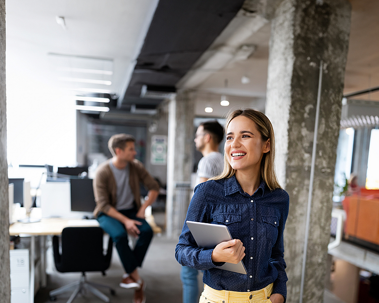 Eine junge Frau lächelt, während sie in einem modernen Büro steht und ein Laptop in der Hand hält. Im Hintergrund sind mehrere Büros und andere Personen zu sehen. Die Frau trägt ein blaues Hemd und gelbe Hosen, was ihr ein frisches Aussehen verleiht.