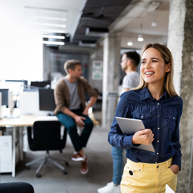 Eine junge Frau lächelt, während sie in einem modernen Büro steht und ein Laptop in der Hand hält. Im Hintergrund sind mehrere Büros und andere Personen zu sehen. Die Frau trägt ein blaues Hemd und gelbe Hosen, was ihr ein frisches Aussehen verleiht.