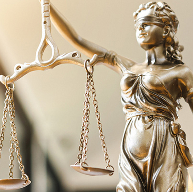 Eine Statue der Justitia, die mit einer Waage in der Hand dargestellt wird. Sie symbolisiert Gerechtigkeit und das Streben nach Fairness im Rechtssystem. Der Hintergrund ist unscharf und zeigt Hinweise auf ein modernes Büro.