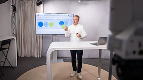 Ein Mann steht an einem Tisch und präsentiert an einem Bildschirm im Hintergrund. Auf dem Bildschirm sind mehrere Farbkreise zu sehen, die möglicherweise ein Diagramm oder eine Grafik darstellen. Der Mann hält einen Stift und erklärt ein Thema.