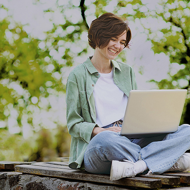 Eine lächelnde junge Frau sitzt im Freien mit gekreuzten Beinen auf einer Holzbank und arbeitet an einem Laptop. Sie trägt eine lässige Jacke und Jeans. Im Hintergrund sind grüne Bäume zu sehen, die eine entspannte und harmonische Atmosphäre schaffen.