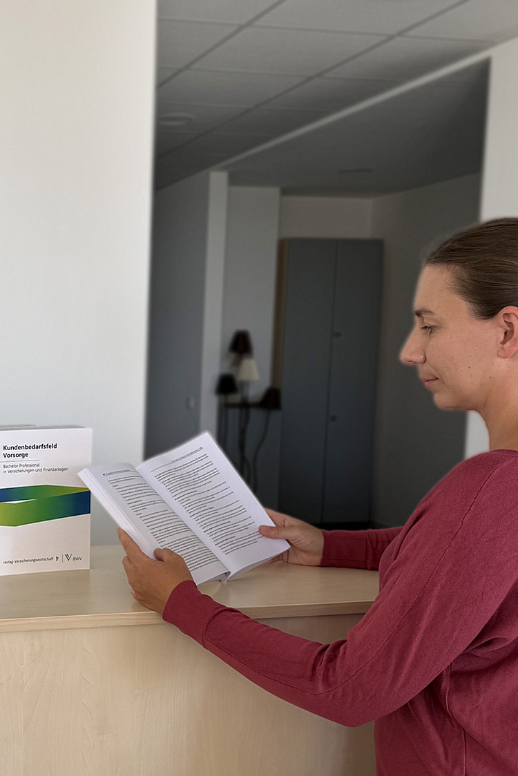 Eine Frau steht an einem Empfangstresen und liest ein Dokument. Auf dem Tresen befinden sich mehrere Broschüren und ein Pflanztopf mit einer Pflanze. Der Raum wirkt hell und modern, mit einer schlichten, neutralen Wandgestaltung im Hintergrund.