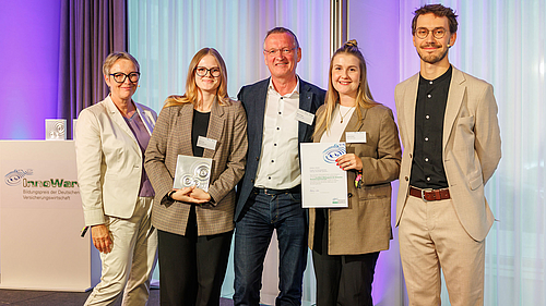 Fünf Personen stehen auf einer Bühne. Zwei Frauen und drei Männer, die alle lächeln. Eine Frau hält ein Zertifikat in der Hand. Im Hintergrund steht ein Tisch mit einem Banner. Die Szene vermittelt ein Gefühl von Auszeichnung und Anerkennung.