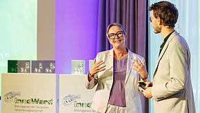 Eine Frau mit Brille steht auf der Bühne und spricht gestikulierend. Neben ihr steht ein Mann im Anzug, der aufmerksam zuhört. Im Hintergrund sind Auszeichnungen auf Podesten sichtbar. Das Bild vermittelt eine Atmosphäre des Dialogs und der Ehrung.