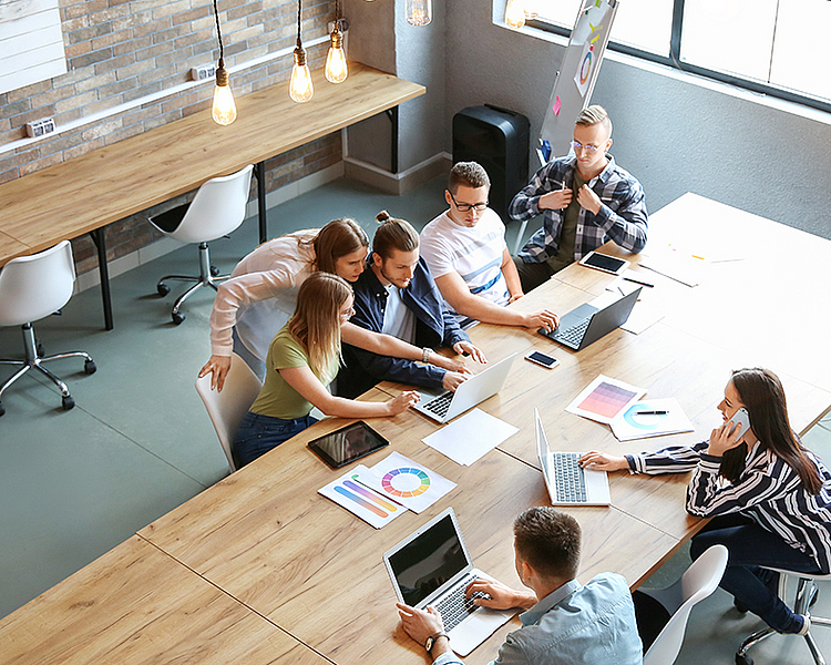 Eine Gruppe von sieben Personen sitzt an einem langen Tisch, arbeitet mit Laptops und diskutiert. Auf dem Tisch liegen Unterlagen und Diagramme. Der Raum ist hell und modern, mit großen Fenstern und minimalistischer Einrichtung.